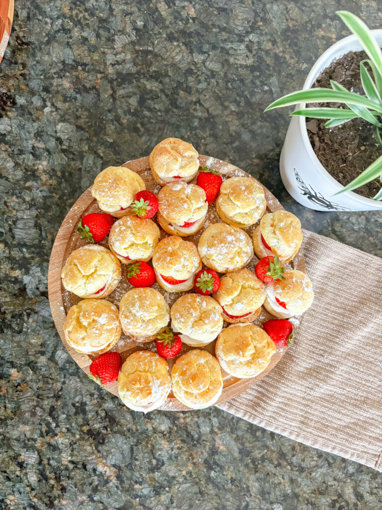 An overhead shot of mini cream puffs resting on a cutting board with strawberries scattered around the board.